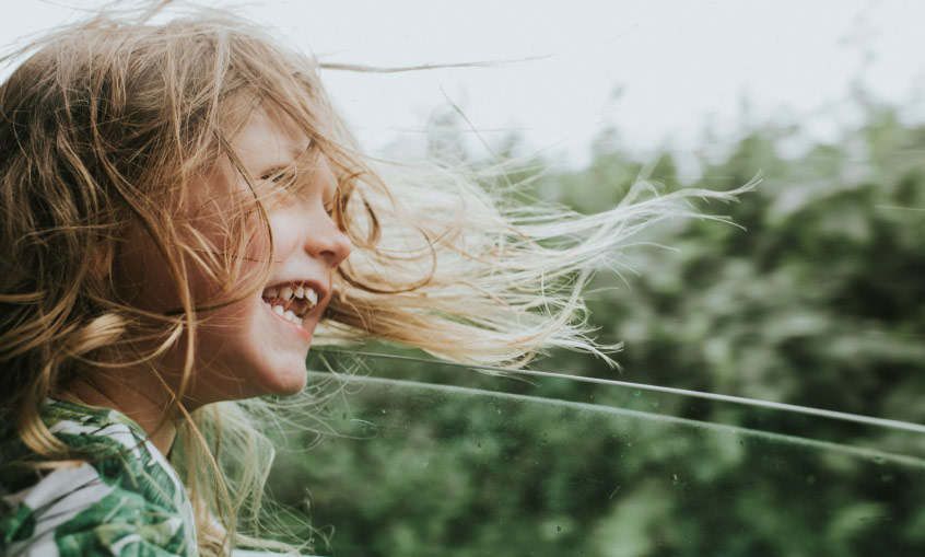 smiling young girl with hair blowing out an open car window driving through a green area